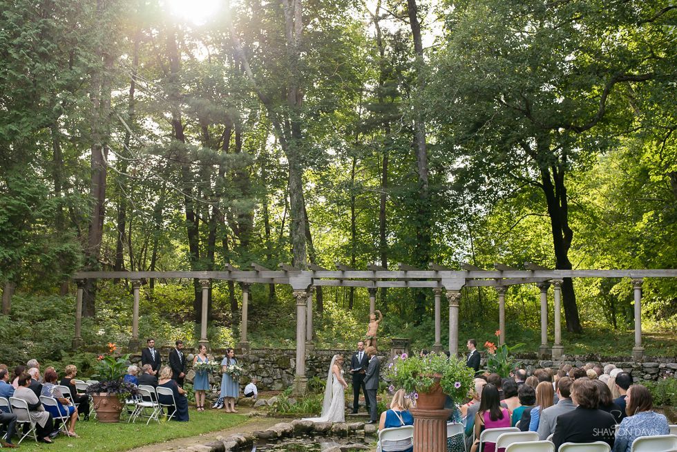 Outdoor wedding ceremony in a sunlit forest clearing, with guests seated beneath a wooden arbor.