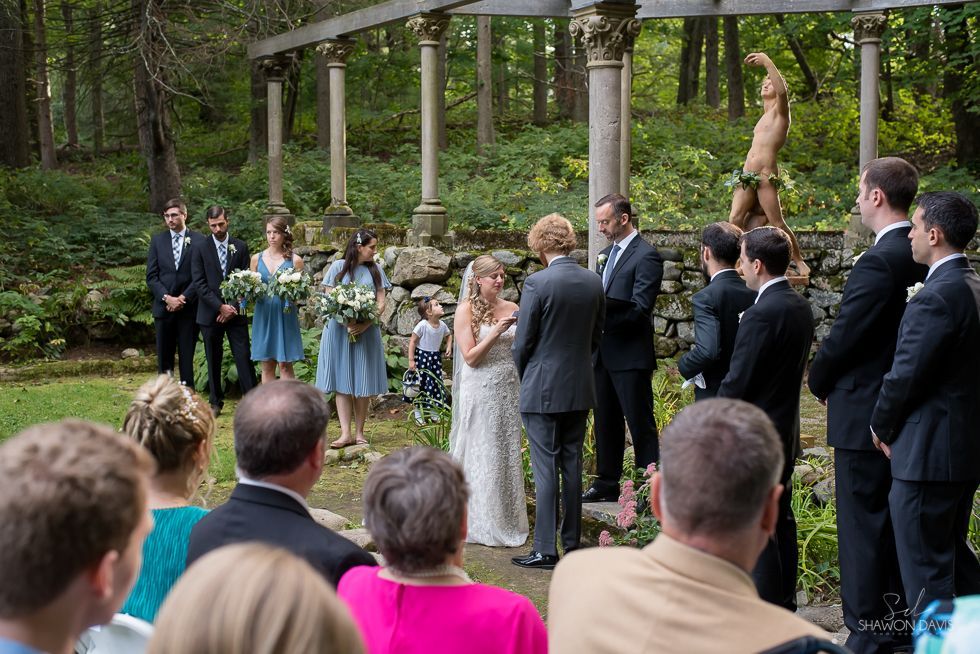 Wedding ceremony outdoors under a stone pergola, with guests seated and the bride and groom standing together.