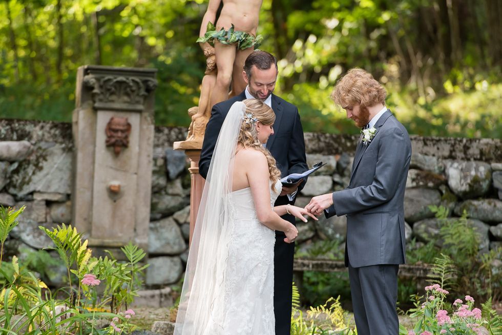 Wedding ceremony outdoors in a garden, couple exchanging rings beside an officiant and floral arch.
