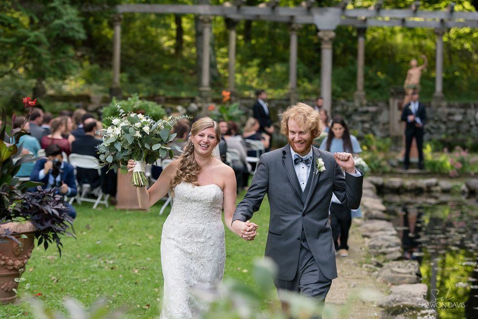 Bride and groom in wedding attire walking hand in hand through a garden ceremony aisle