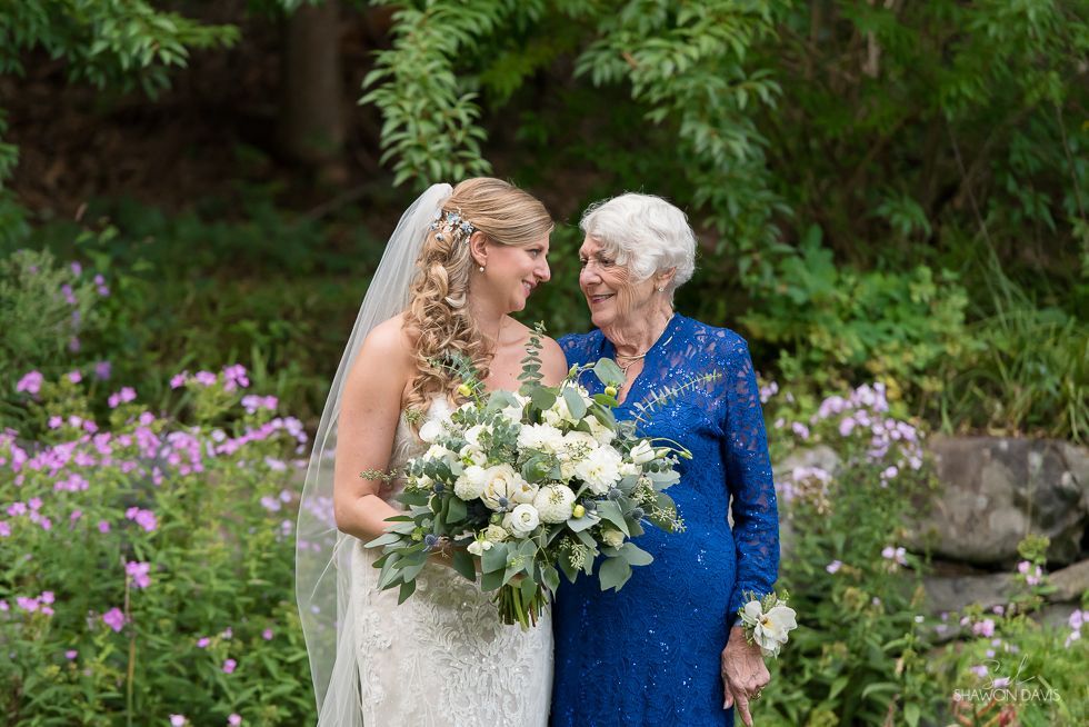 Bride in white dress and elderly woman in blue dress holding flowers in a garden.