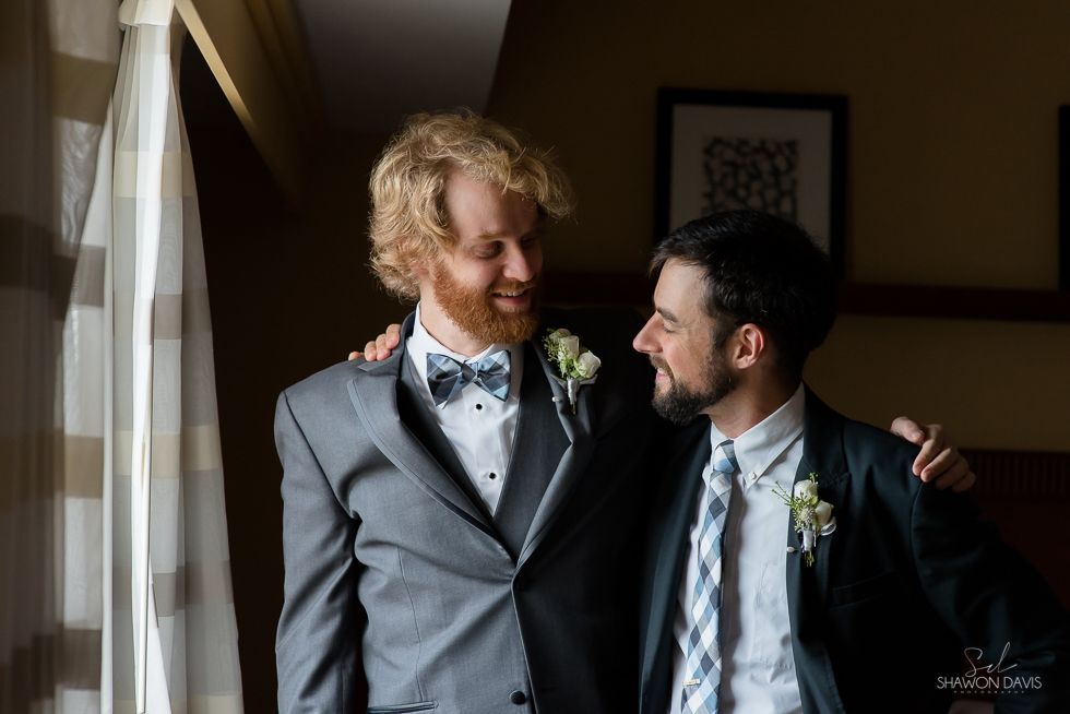 Two grooms in suits smiling at each other by a window, one with an arm around the other.