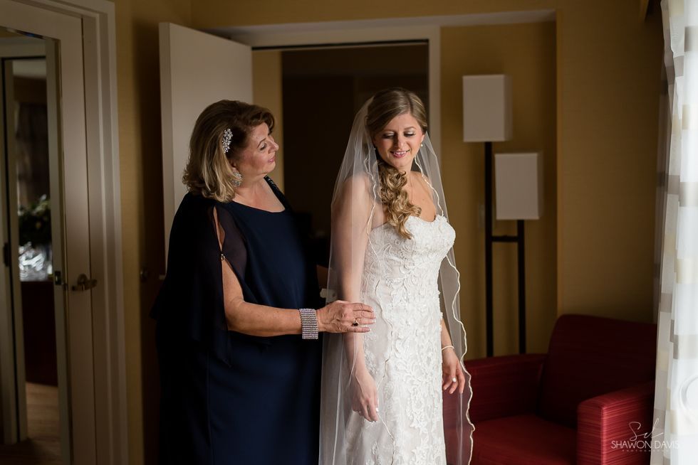 Woman in a white wedding dress stands with an older woman in a dark dress indoors.