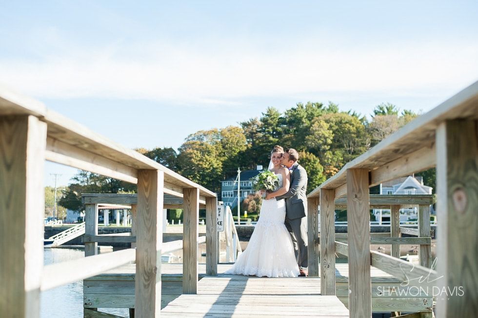 Bride and groom standing on a wooden dock, embracing in bright daylight by the water