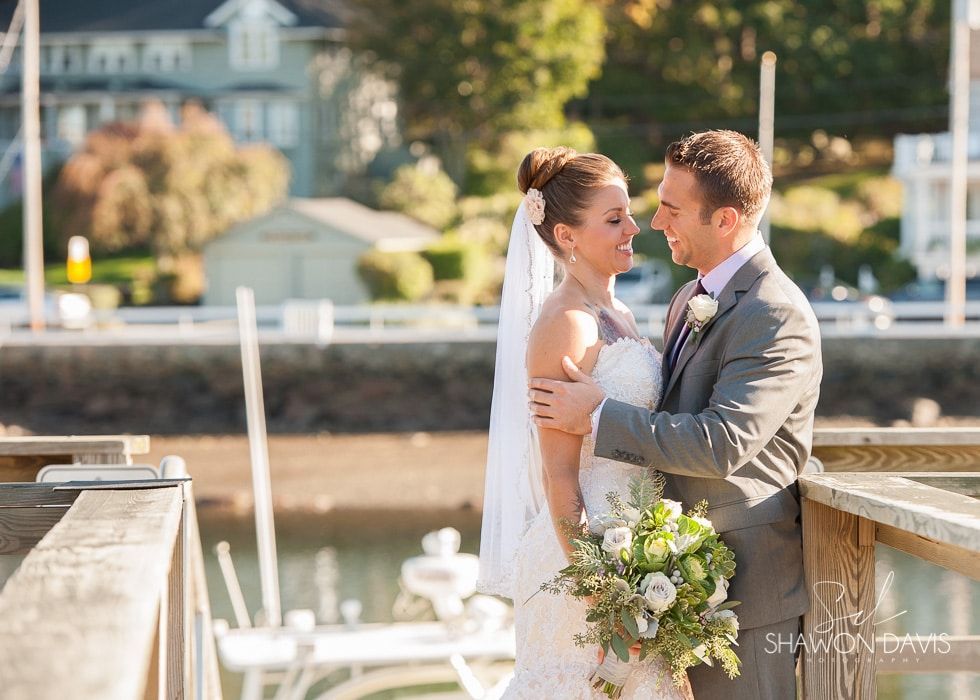 Bride and groom embracing by a waterfront dock, with a bouquet and wedding attire in soft sunlight.