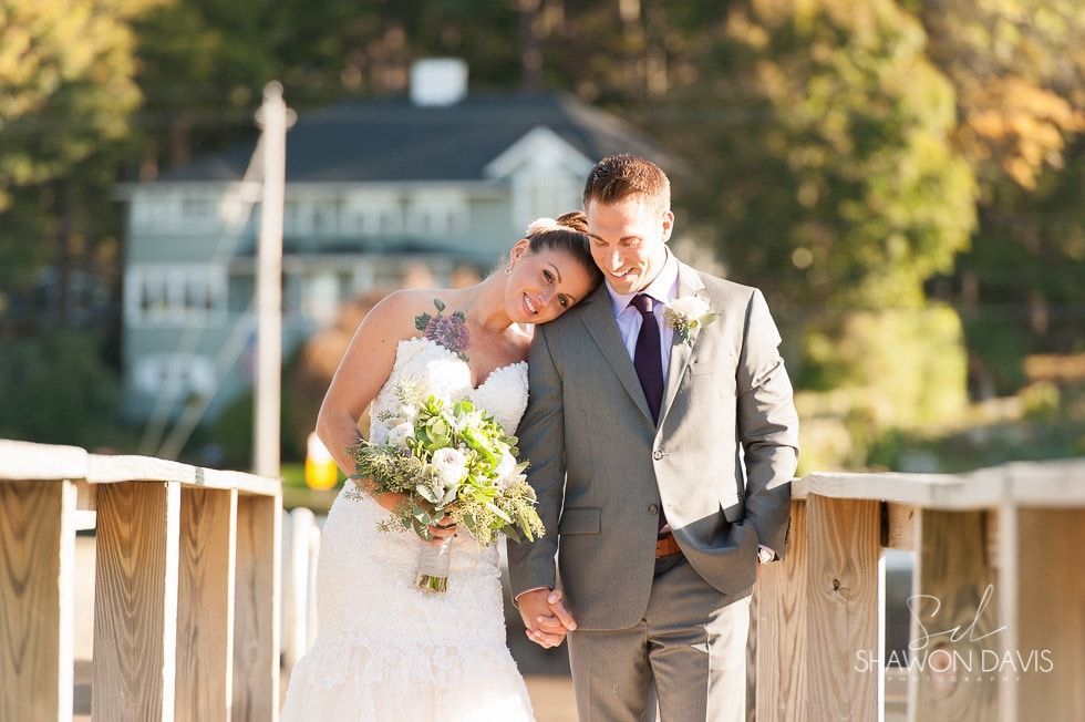 Bride leaning on groom’s shoulder on a sunlit wooden bridge, holding a bouquet.