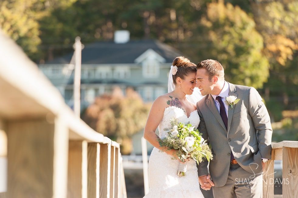 Bride and groom touching foreheads on a sunlit bridge, holding a bouquet, with a blurred house in the background.