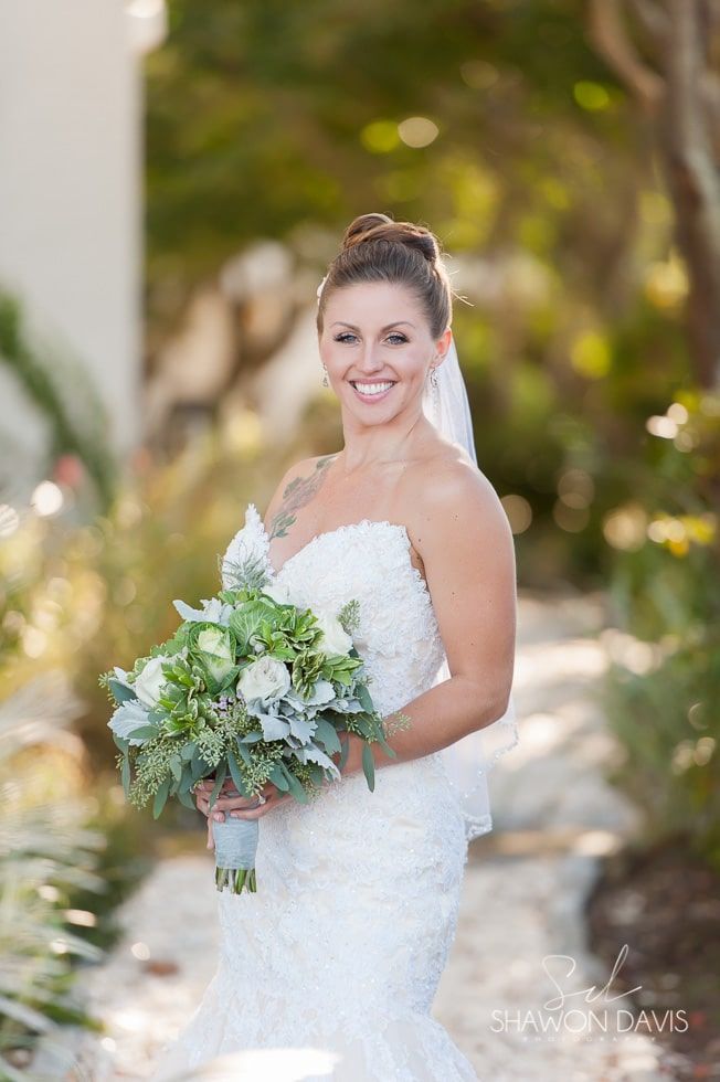 Bride in strapless white gown holding a bouquet, smiling outdoors with a blurred garden background