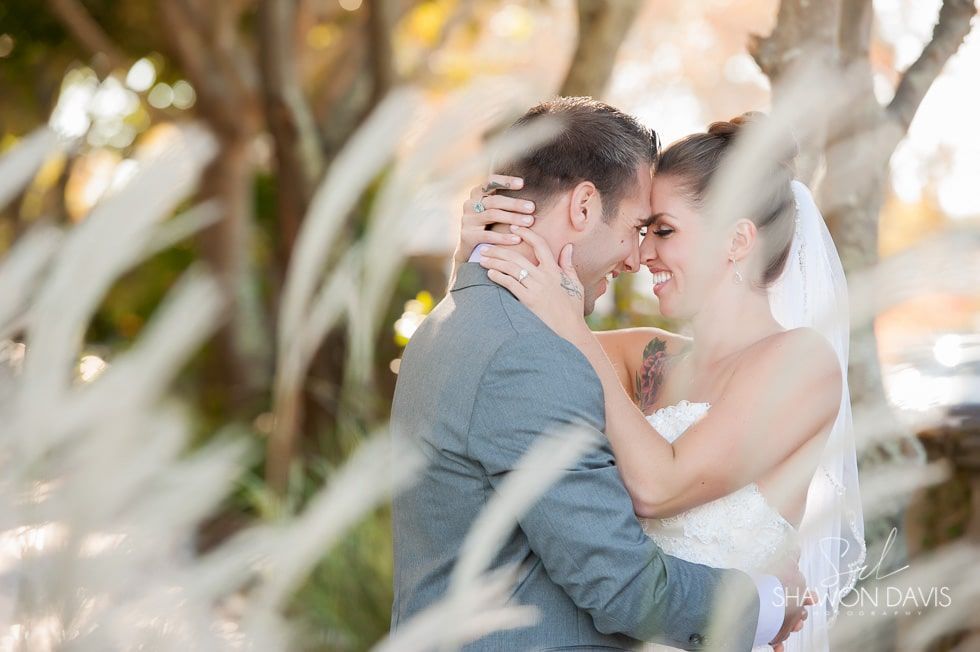 Bride and groom embrace and smile outdoors, framed by soft white foliage.