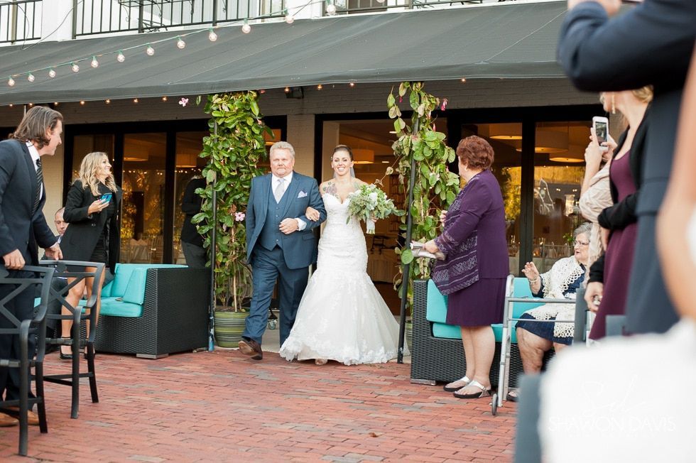 Bride and groom exit a venue doorway as guests watch and take photos on a brick patio.