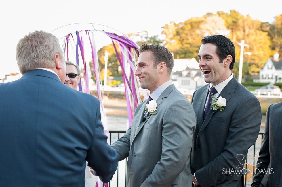 Two grooms in gray suits smile during an outdoor wedding ceremony by a colorful fountain.