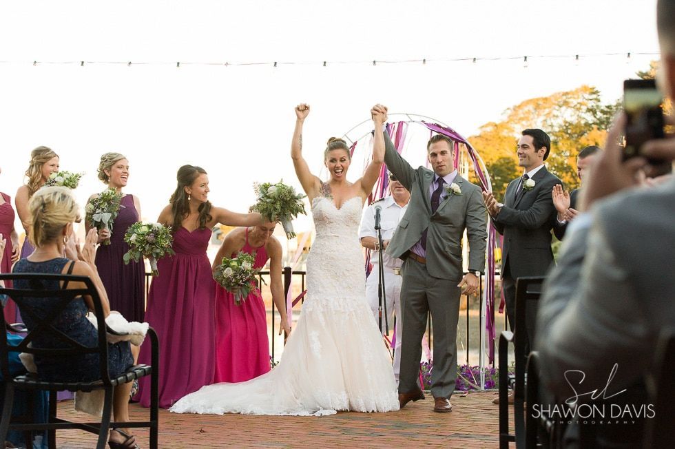 Wedding ceremony with couple raising hands, guests cheering, and bridesmaids in purple beside an officiant outdoors