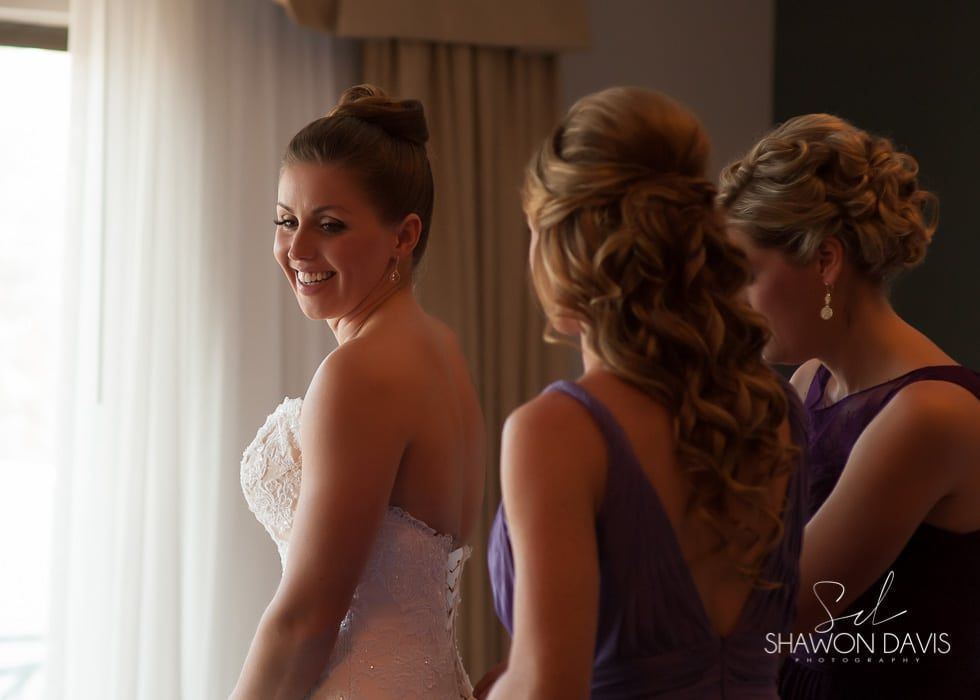 Bride smiling in a strapless white gown, with two bridesmaids in purple dresses nearby by a window