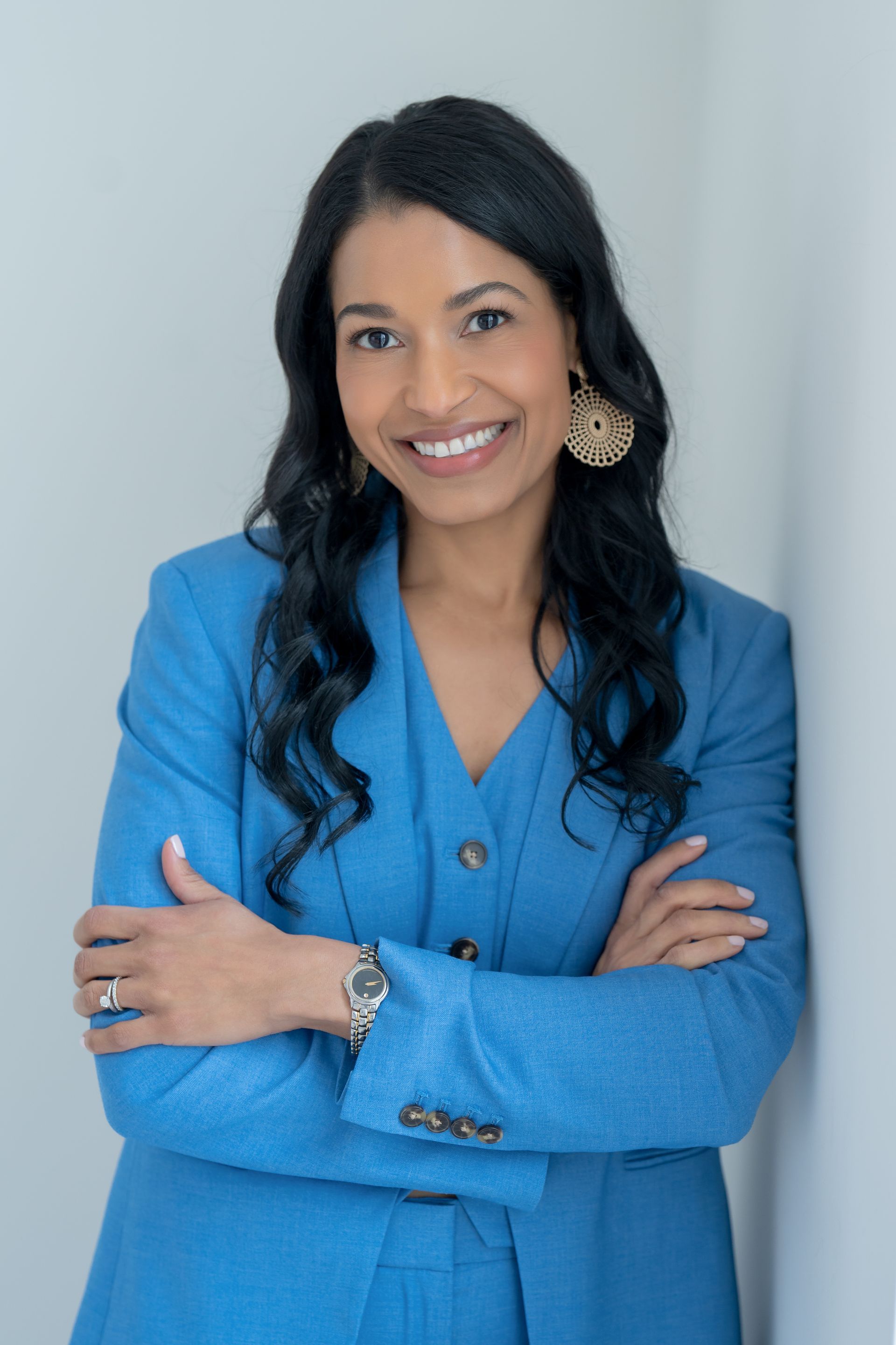 Woman in a three-piece blue suit smiling while standing against a light wall.