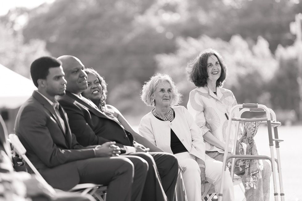 Seated guests in formal attire listen outdoors at a wedding ceremony
