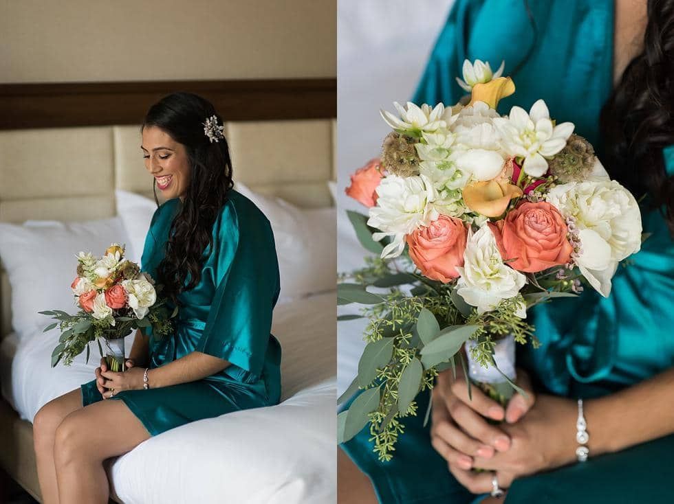 Woman in a teal dress sitting on a bed, smiling and holding a bouquet of peach and white flowers