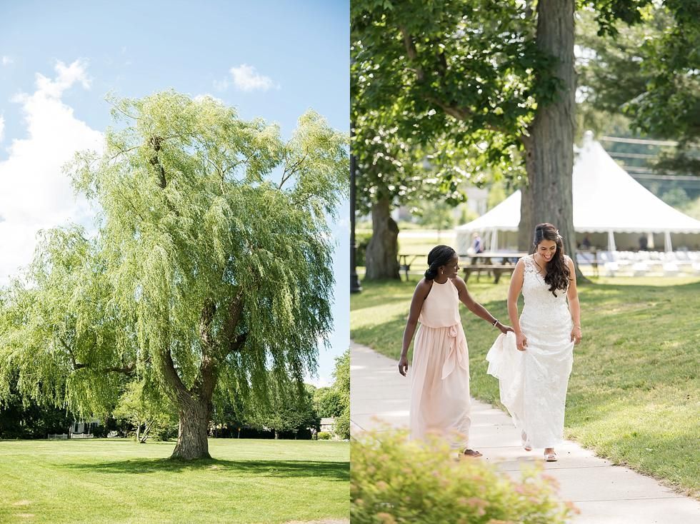Two women in light dresses walking hand in hand on a sunny path beside a white tent and trees.