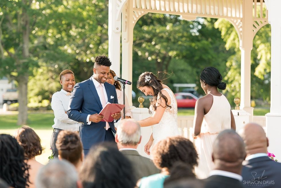 Outdoor wedding ceremony under a gazebo, couple exchanging vows as officiant reads from a book, guests seated nearby