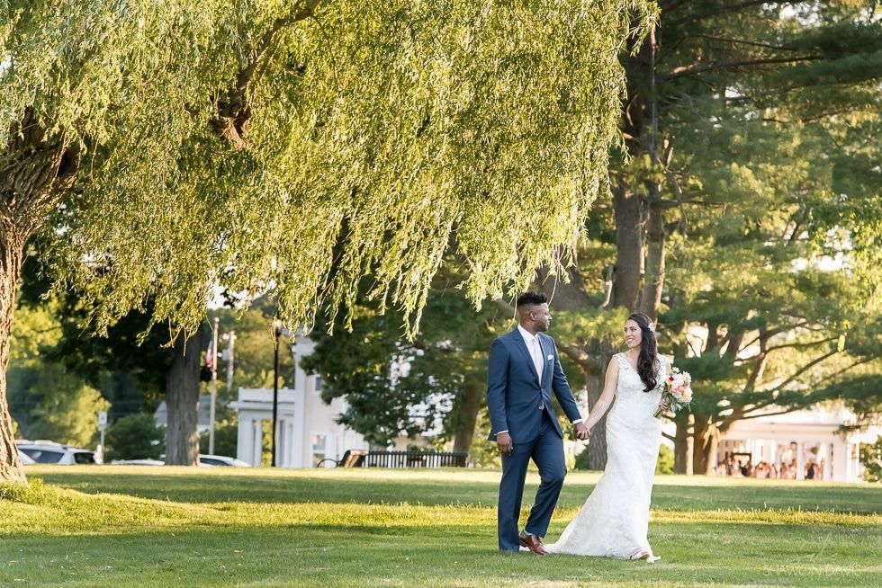 Bride and groom walk hand in hand on a sunny lawn under a large tree