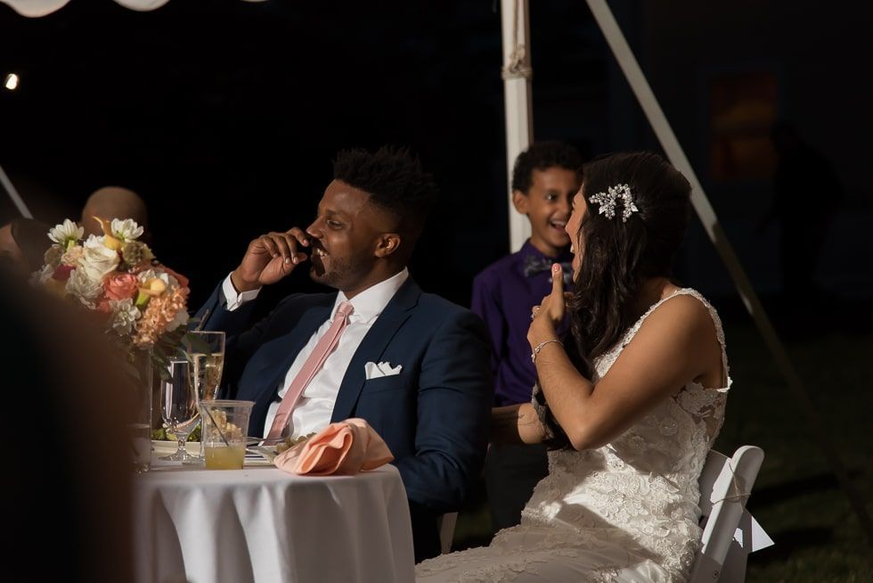 Bride and groom seated at a candlelit wedding reception table, with a man speaking in the background