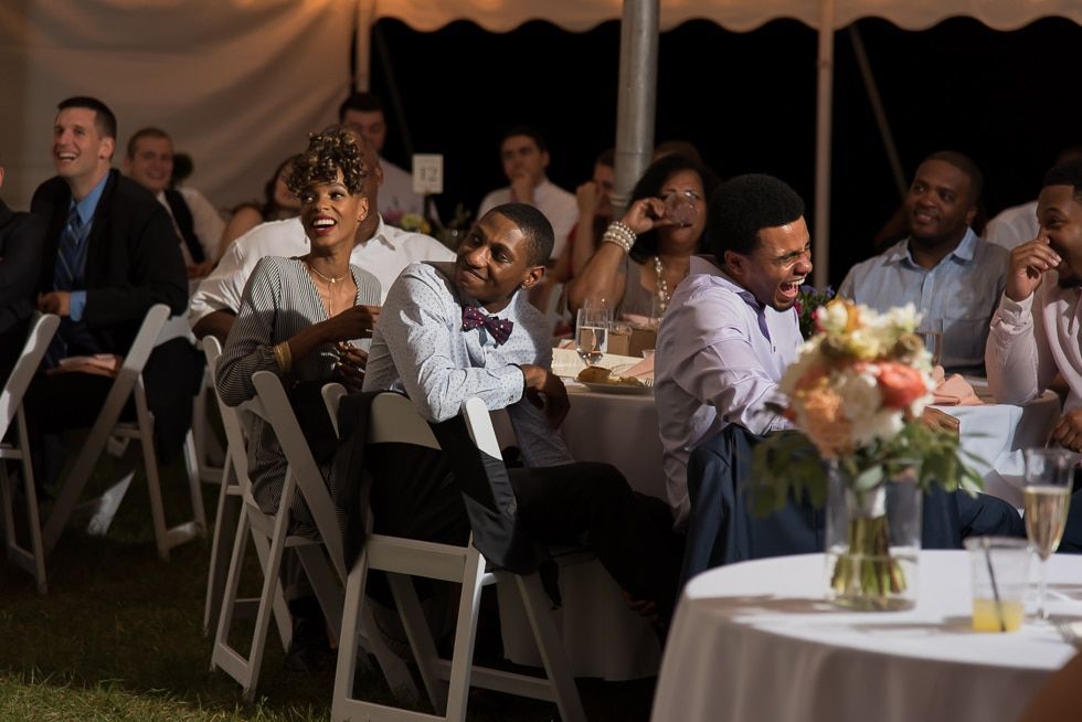 Guests seated at a tented event, smiling and looking toward the camera, with floral table centerpiece