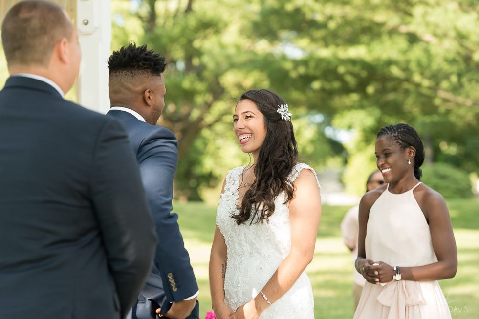 Bride smiling at an outdoor wedding ceremony, surrounded by guests and a suited officiant.