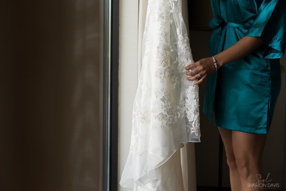 Person in a teal dress adjusts a white lace wedding gown by a window.