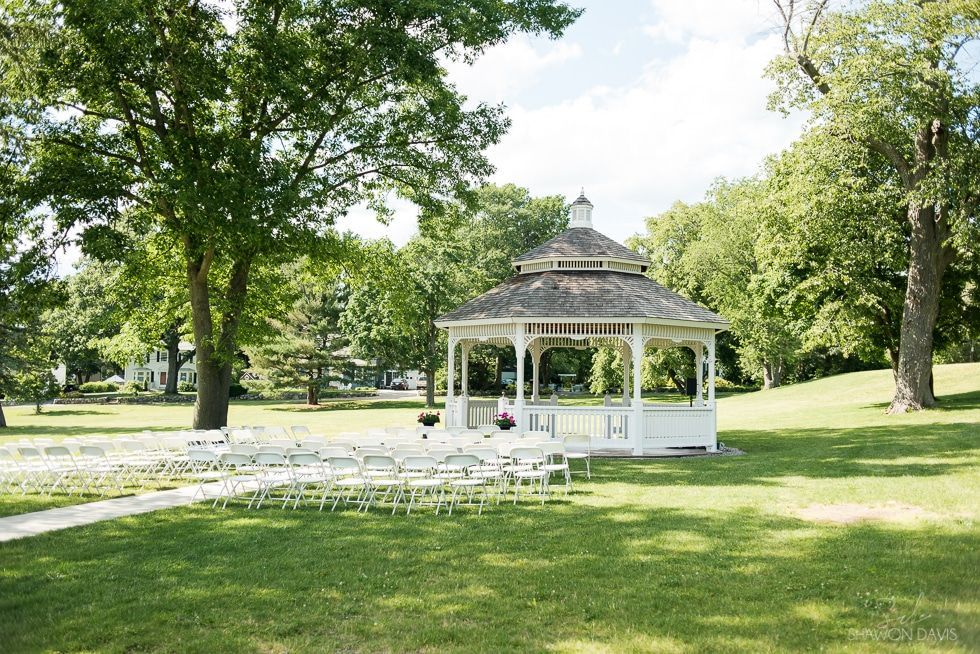 White gazebo in a sunny park with green lawn and trees, surrounded by rows of chairs