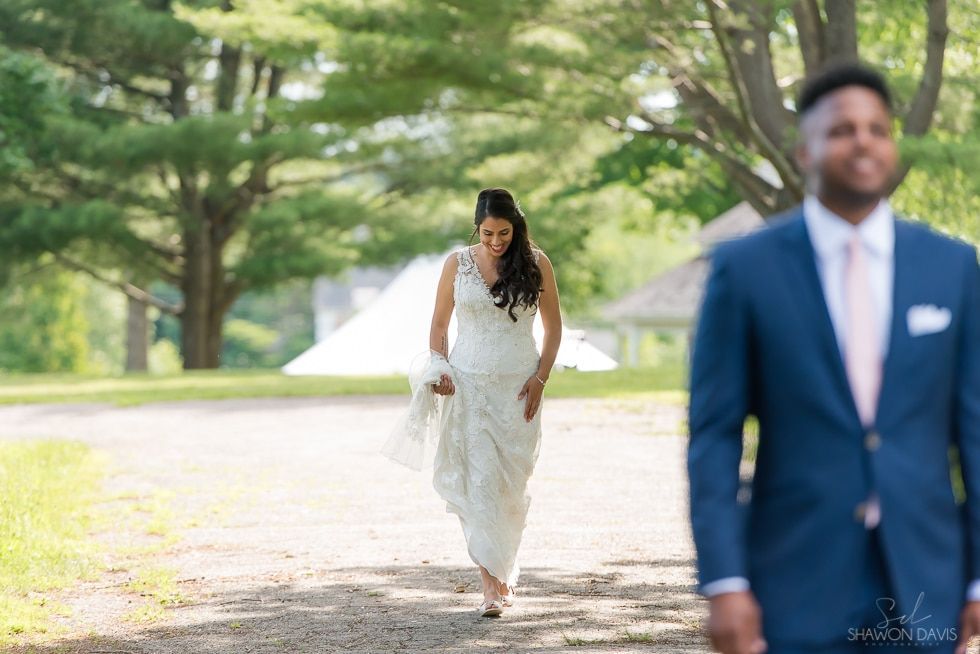 Bride walking down a sunlit path toward groom in blue suit, with trees and a house in the background