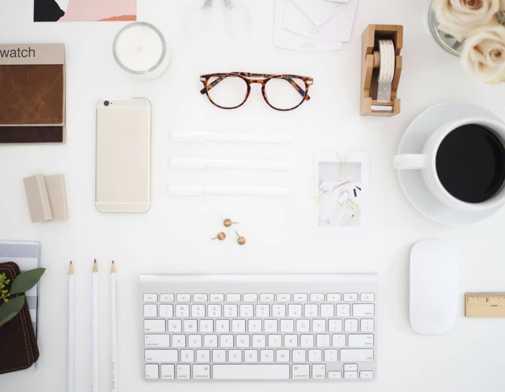 Minimal white desk with keyboard, coffee, glasses, phone, clock, and stationery arranged neatly from above