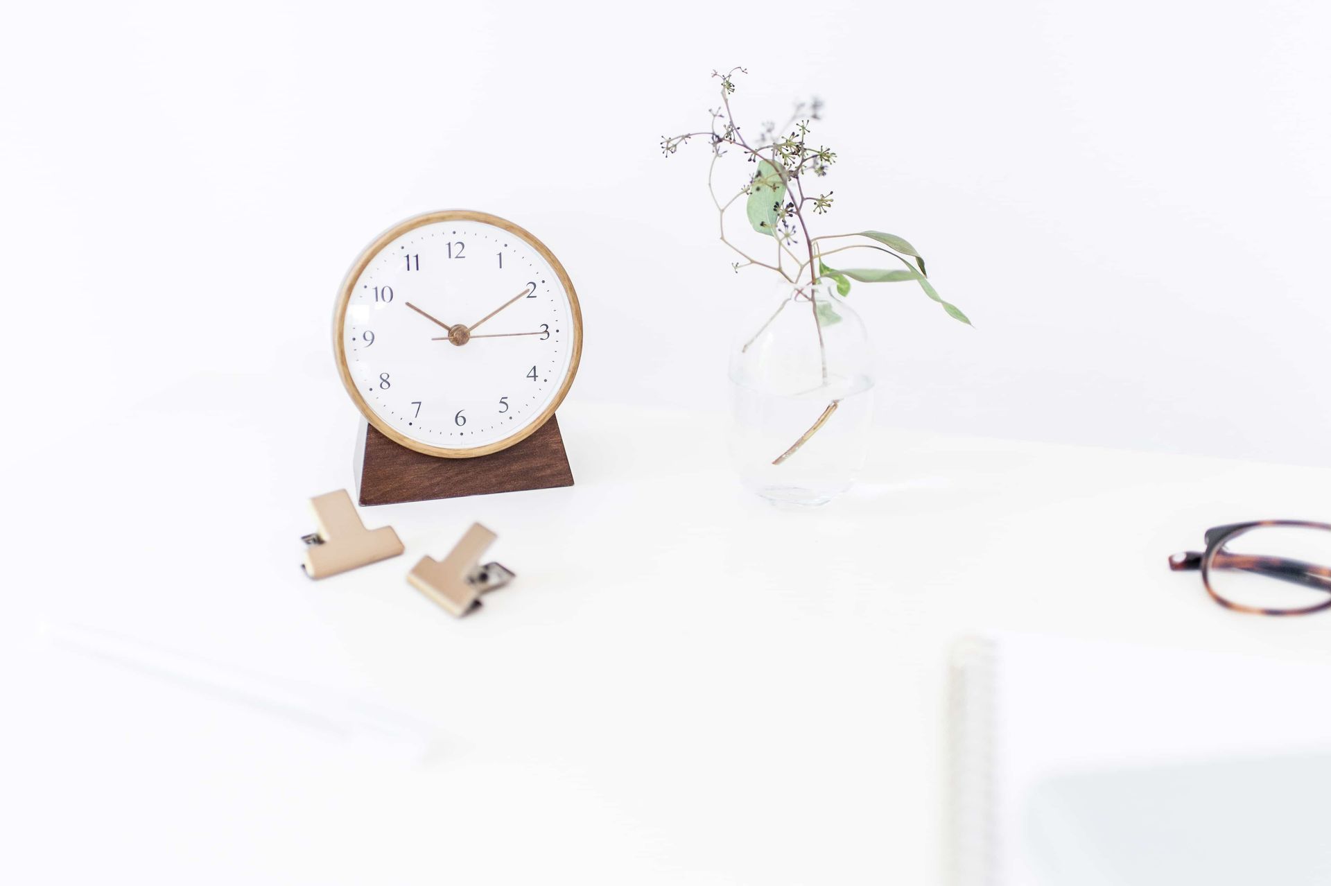 Minimal desk with a wooden clock, small plant, and eyeglasses on a white surface.