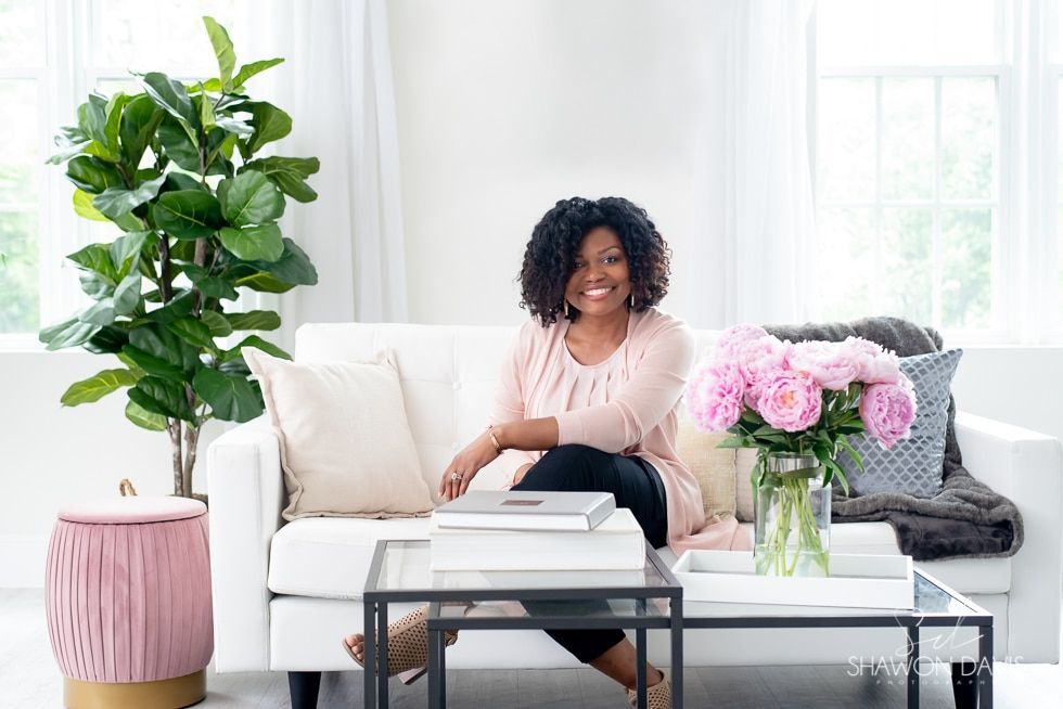 Woman seated on a white sofa in a bright living room with pink flowers, a coffee table, and a potted plant.