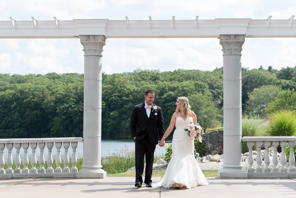 Bride and groom standing under white columns by a lake, holding hands on a sunny day