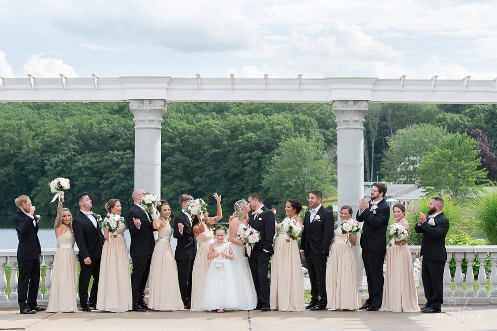 Wedding party posing outdoors under a white pergola, with white and black formal attire and bouquets.