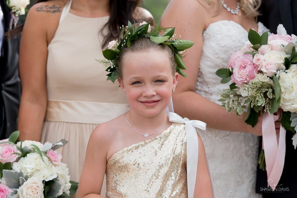 Young child in a gold dress with green leaf crown, standing beside bridesmaids holding pink bouquets.