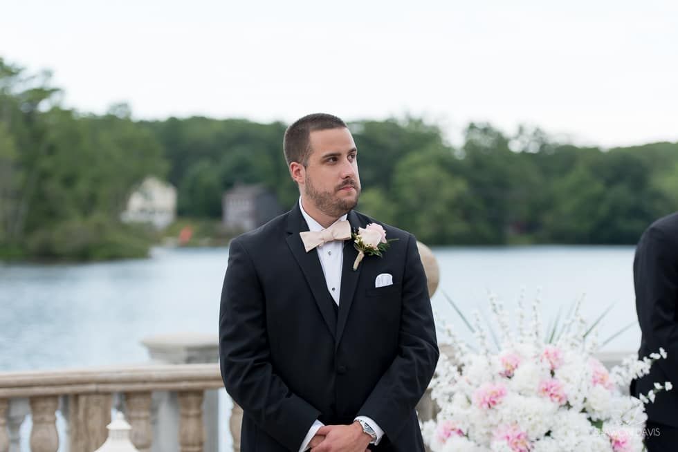 Man in a black tuxedo standing outdoors by the water at a wedding ceremony