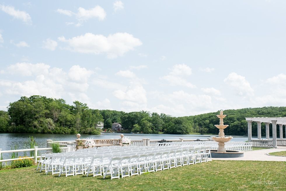 White chairs on a lakeside lawn beside a fountain under a partly cloudy sky