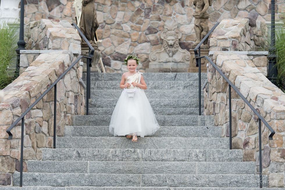 Bride in white dress holding bouquet on stone staircase, framed by rustic stone walls.