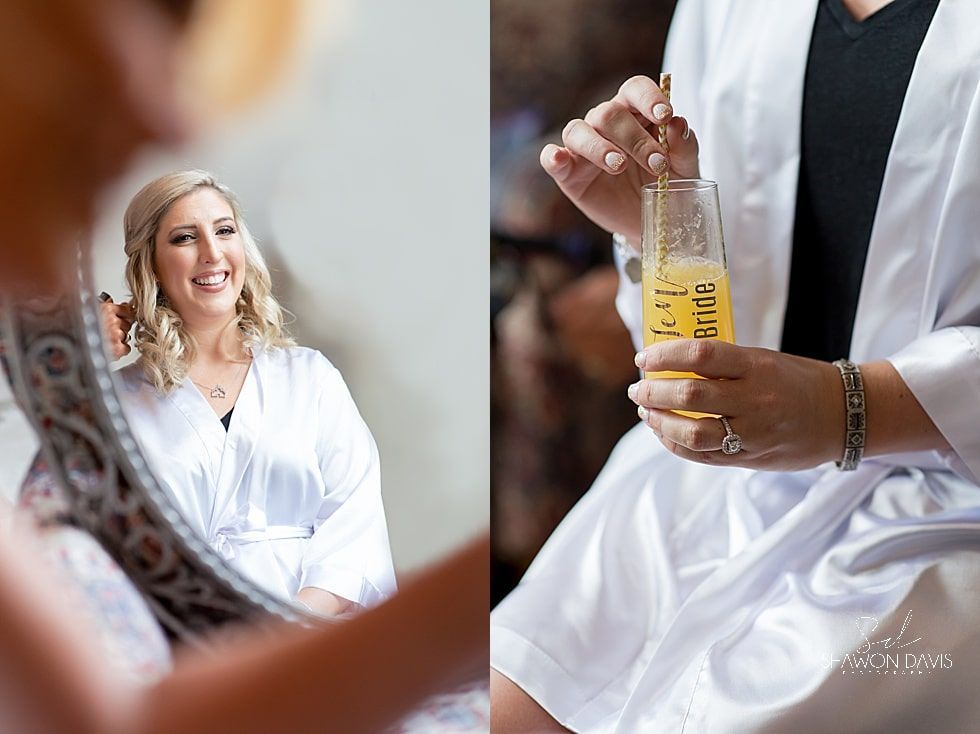 Bridal party detail: hands opening a champagne bottle in white robes, with a smiling woman reflected in a mirror