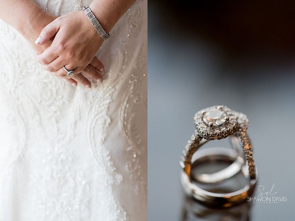 Close-up of a bride’s hand with diamond rings beside a white wedding dress and stacked rings