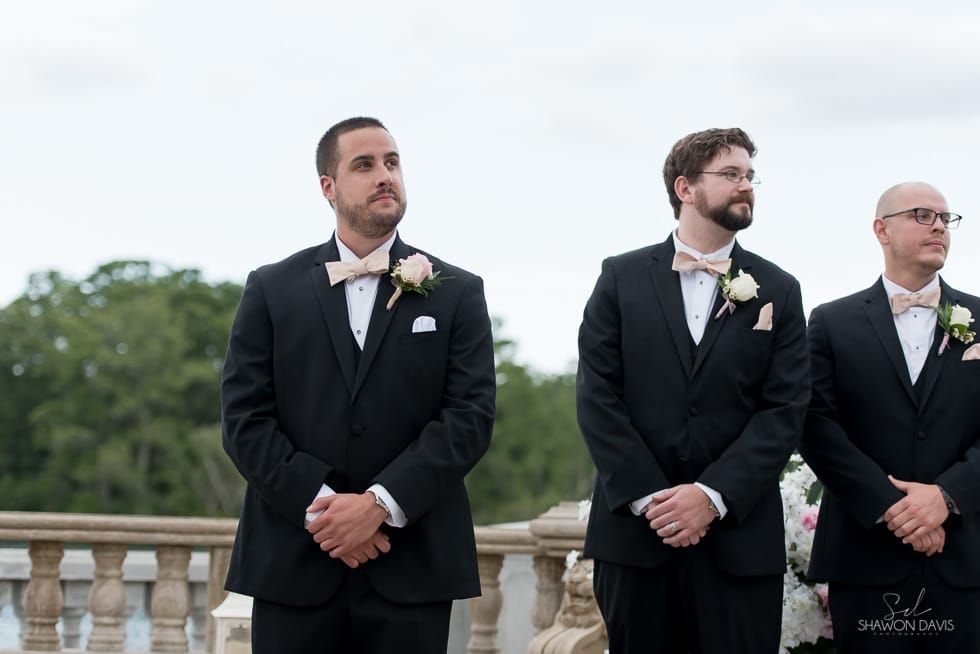 Three men in black tuxedos stand outdoors, waiting at a wedding ceremony.