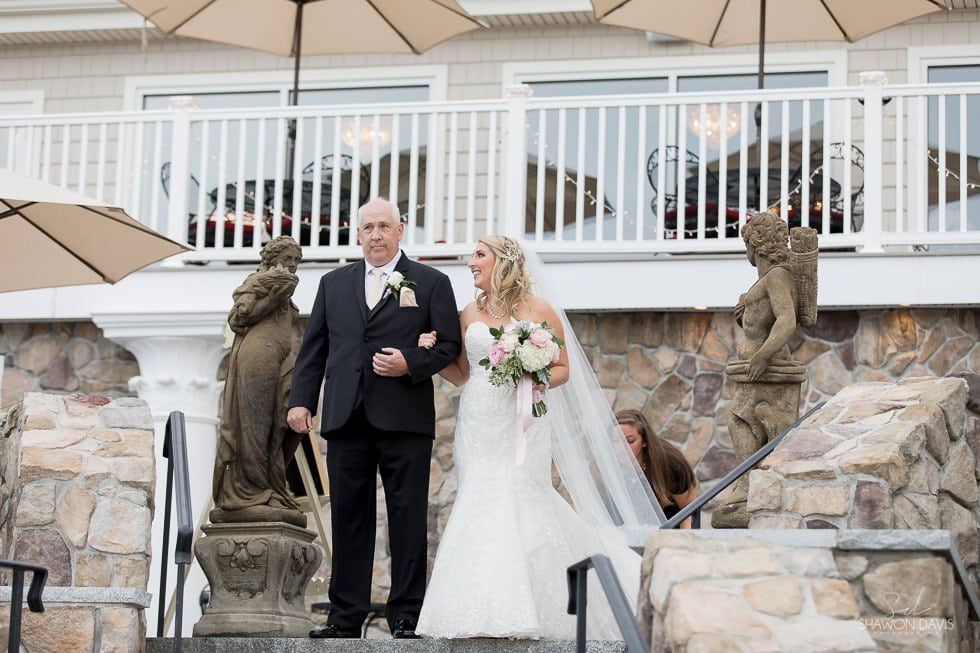 Bride and groom descend stone steps at an outdoor wedding venue, flanked by statues and white railings.