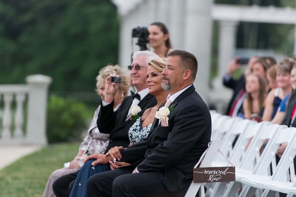 Guests seated at an outdoor wedding ceremony with a groom in a black suit in the foreground