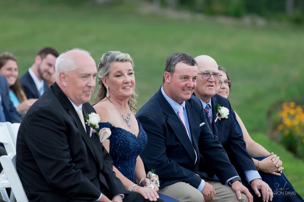 Guests seated outdoors at a wedding ceremony, dressed in formal attire and smiling