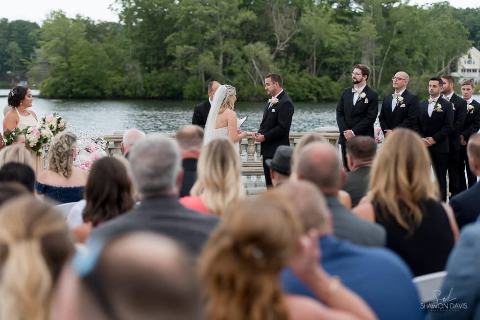 Outdoor wedding ceremony by a lake, with bride, groom, and attendants facing seated guests.