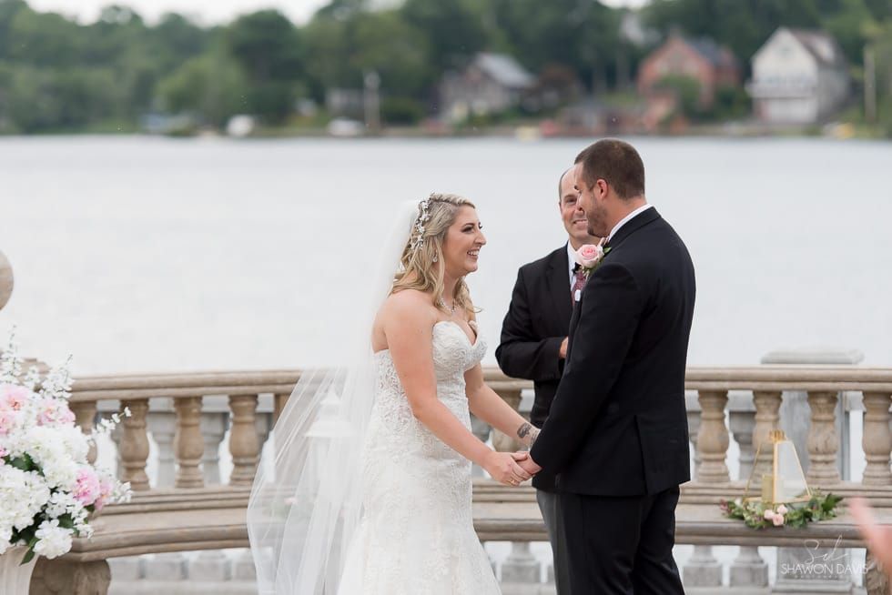 Bride and groom exchanging vows by a lakeside railing with an officiant nearby