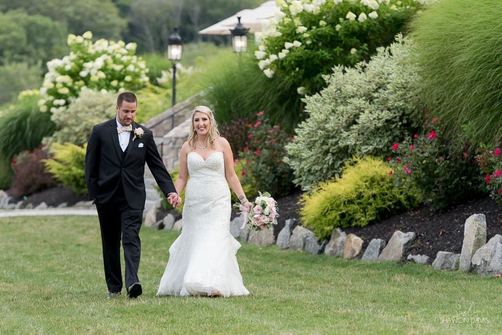 Bride and groom walking on a grassy path in a garden with white flowers and stone landscaping