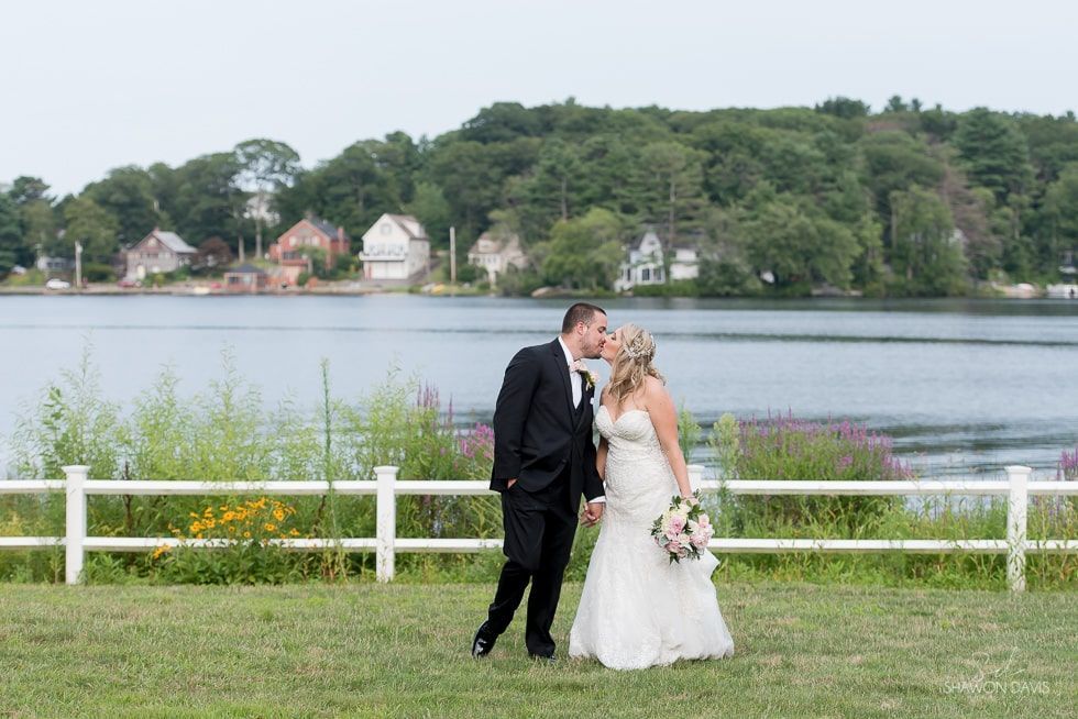 Bride and groom kissing by a lakeside fence with trees and houses across the water