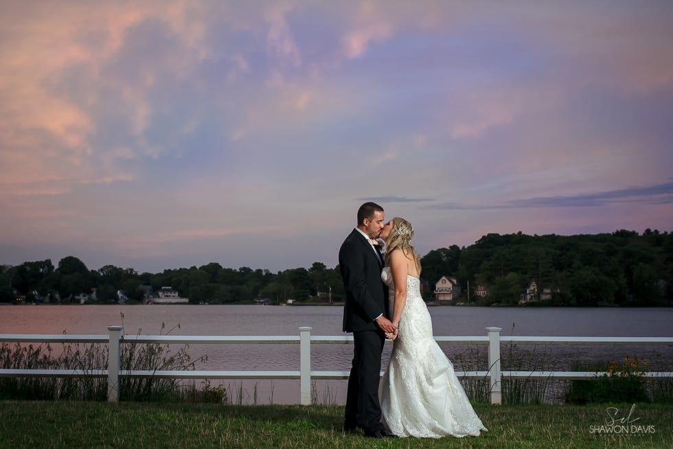 Bride and groom embrace by a lakeside fence at sunset under a pastel sky.