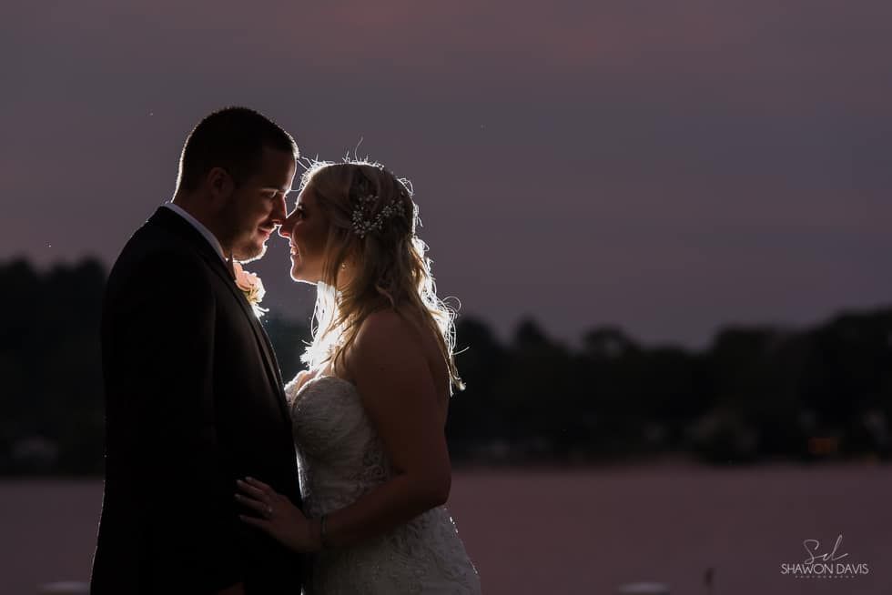 Silhouetted couple embracing at dusk by the water, with soft backlighting and a blurred horizon.